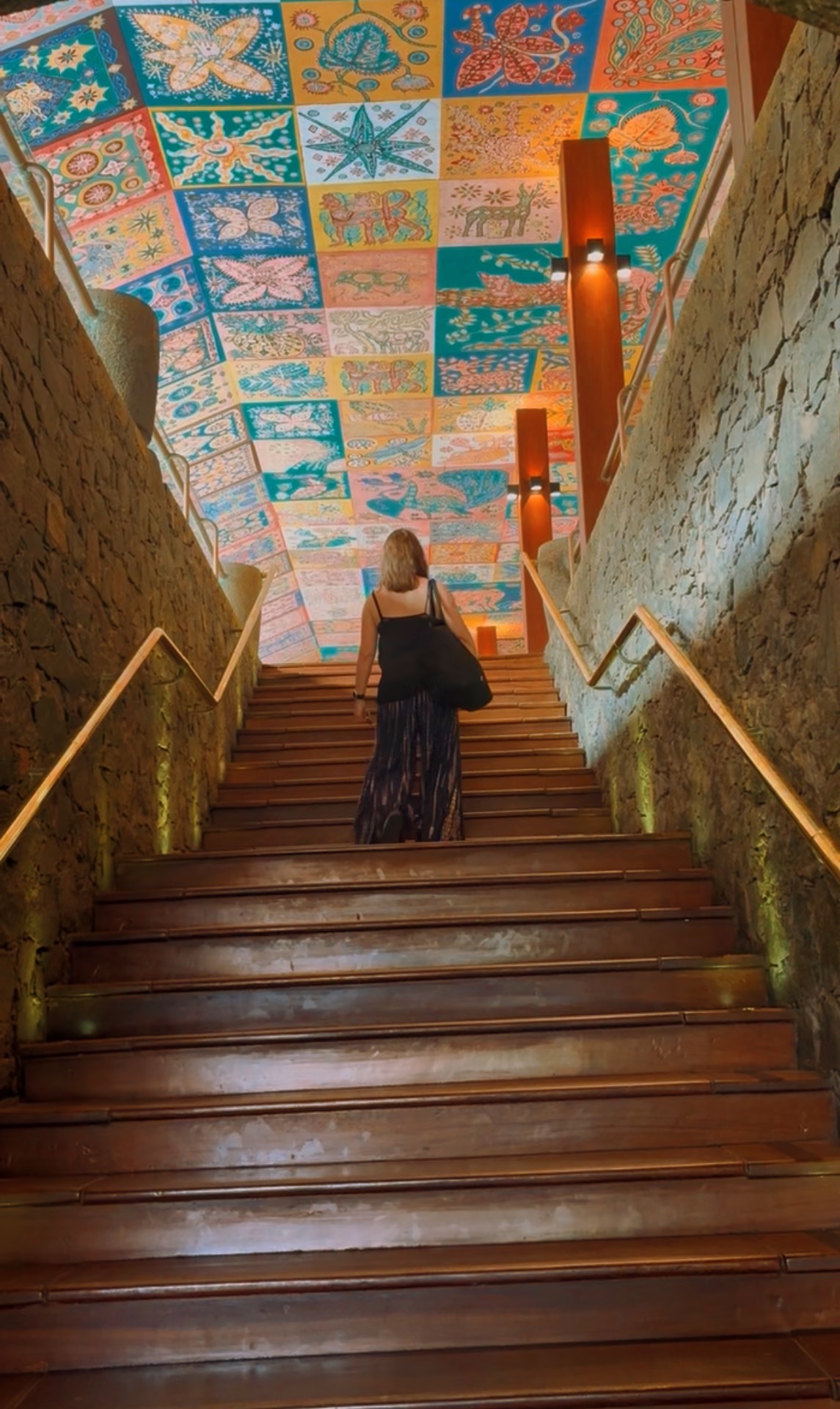 Woman walking up wooden stairs beneath a colourful batik ceiling mural
