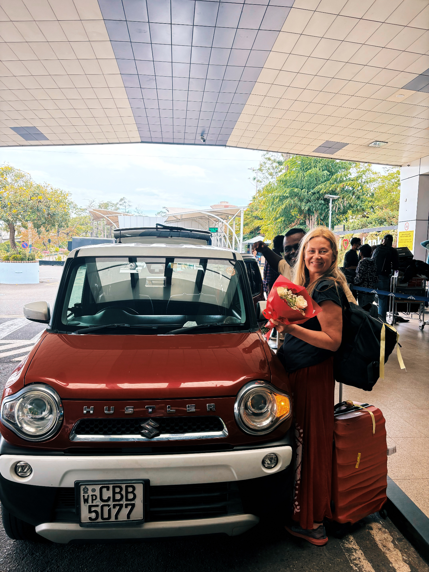 Retreat guest welcomed at the airport beside a red Suzuki Hustler with flowers and luggage
